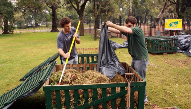 Dos hombres removiendo y vaciando residuos orgánicos en una compostera grande de madera verde en un parque.