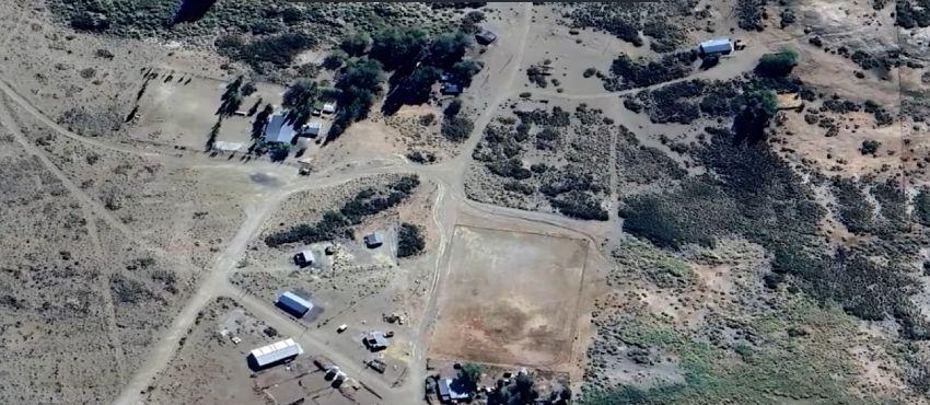 Vista aérea o satelital de la comunidad Millaqueo, que muestra un terreno rural árido con caminos de tierra, algunas construcciones y viviendas dispersas, vegetación baja, árboles agrupados y una cancha o terreno rectangular delimitado.