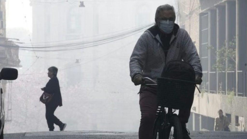Persona en bicicleta y peatones circulan por una calle urbana cubierta de smog y contaminación del aire