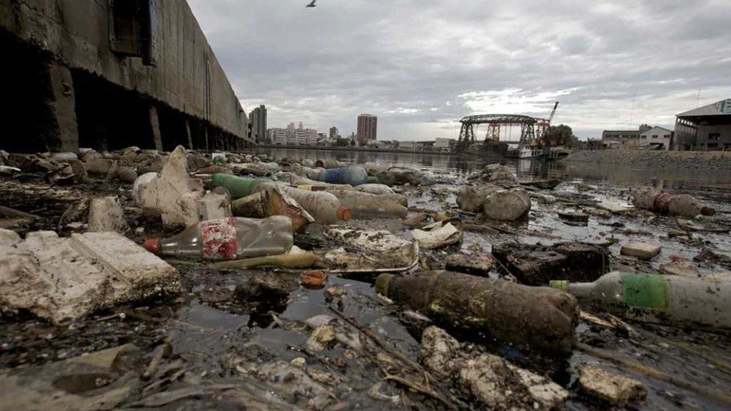 Residuos plásticos y otros desechos acumulados en la orilla de un curso de agua contaminado, junto a estructuras industriales y urbanas