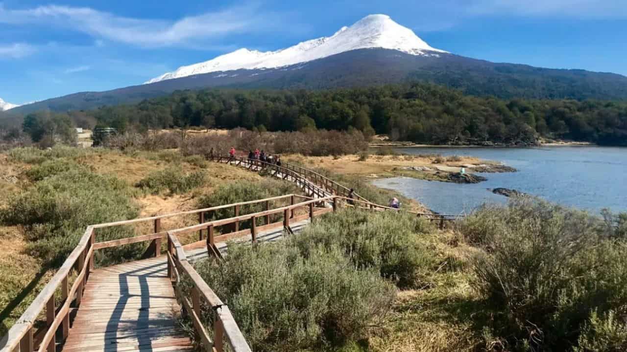 Sendero de madera junto a un lago en el Parque Nacional Tierra del Fuego, con vegetación nativa y montañas nevadas de fondo
