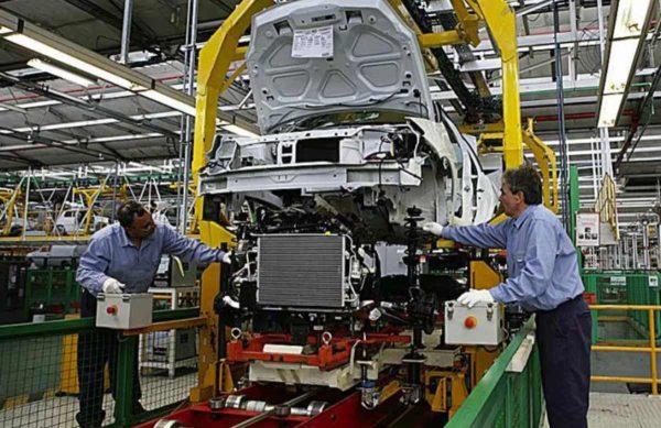 Dos operarios ensamblan la parte frontal y el radiador de la carrocería de un automóvil blanco suspendido en la línea de montaje de una fábrica automotriz.