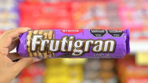 Una mano sosteniendo horizontalmente un paquete violeta de galletitas Frutigran con chips de chocolate de la marca Granix. En la parte superior derecha del envase se aprecian los sellos negros de exceso en azúcares y calorías, con un fondo de góndolas de supermercado desenfocado.