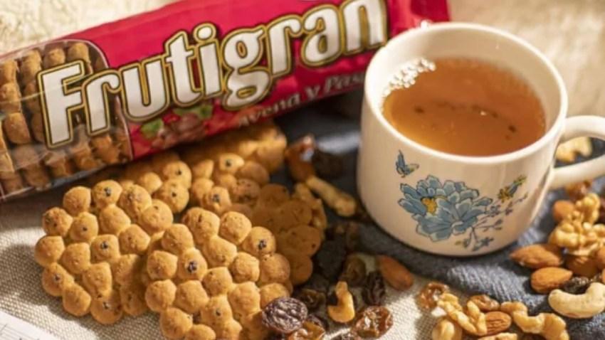 Un paquete rojo de galletitas Frutigran sabor avena y pasas al lado de una taza de té blanca con detalles florales azules. En primer plano hay varias galletitas redondas de avena acompañadas por pasas de uva y frutos secos variados como nueces y almendras sobre un mantel claro