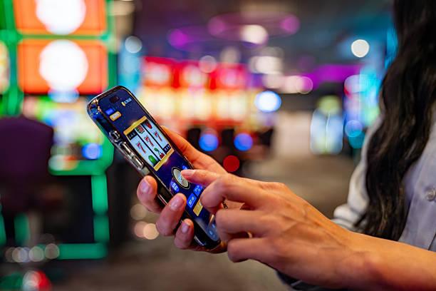 Primer plano de una persona jugando a la tragamonedas desde un teléfono inteligente en el interior de un casino iluminado.