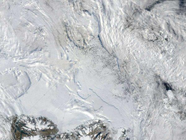 Imagen satelital de la región del Océano Ártico que muestra extensas capas de hielo marino y cobertura de nubes, con líneas de latitud y longitud convergiendo cerca del Polo Norte. La parte inferior de la imagen muestra la costa ártica, con masas de tierra rocosas cubiertas de nieve que delimitan las aguas heladas.