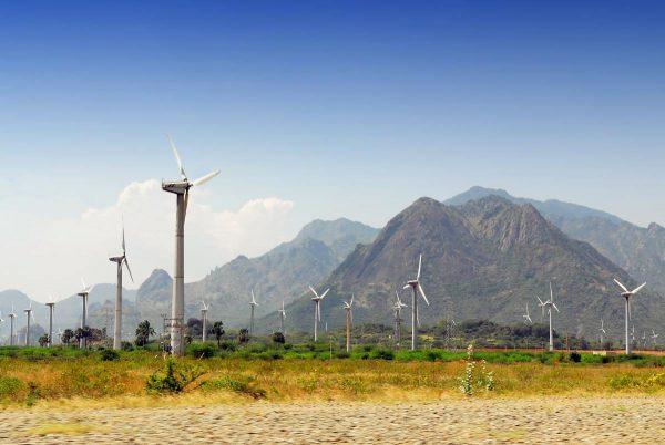 Parque eólico con múltiples aerogeneradores distribuidos en un llano, con colinas y montañas verdes al fondo bajo un cielo despejado.