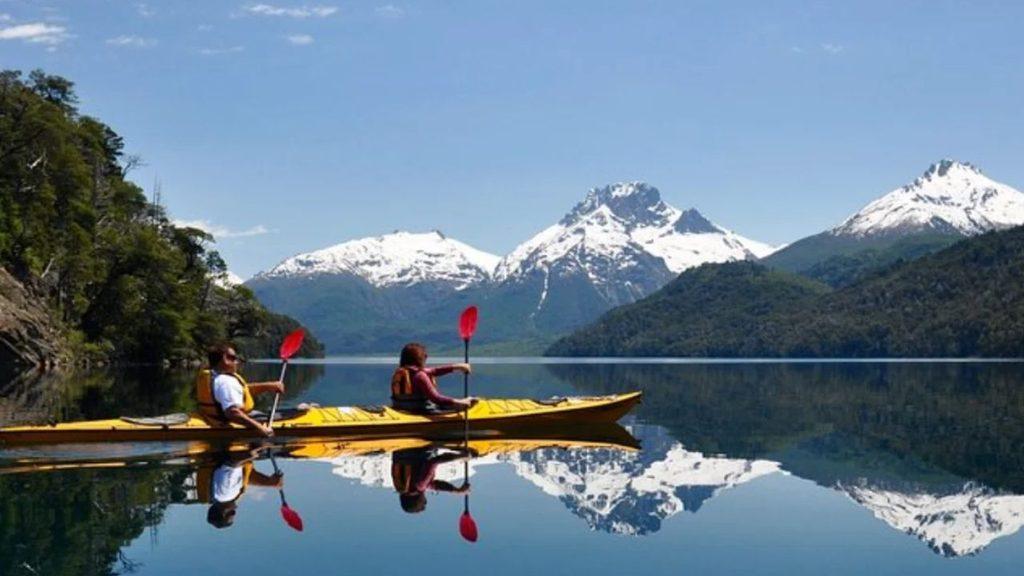 Dos personas practican kayak en un lago patagónico de aguas tranquilas, rodeado de bosques y montañas nevadas