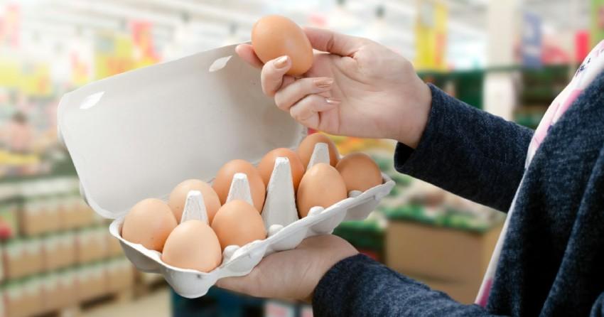Una persona sosteniendo una caja de cartón abierta con huevos marrones en un supermercado, mientras toma un huevo con la mano derecha.