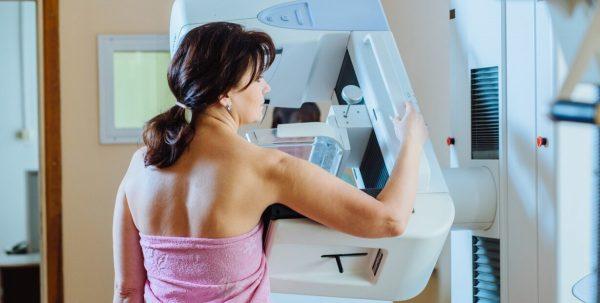 Una mujer de espaldas, envuelta en una toalla rosa, acomodándose para realizarse un estudio médico en un mamógrafo blanco.
