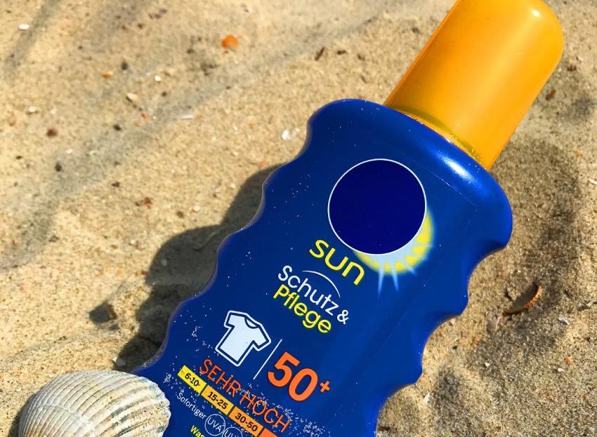 Botella azul de protector solar SPF 50+ colocada sobre la arena de una playa junto a una concha marina