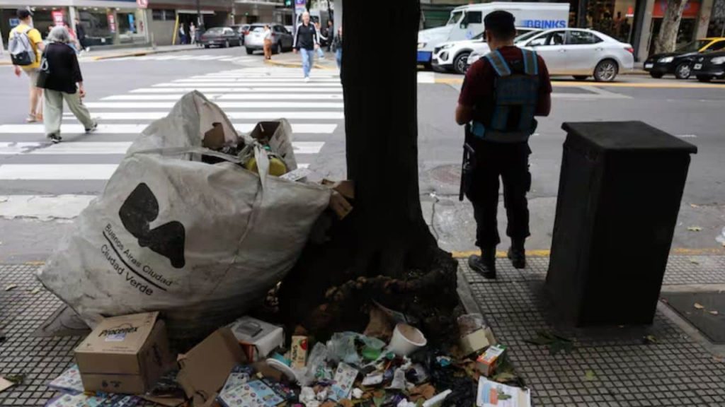una bolsa de basura utiliazada por cartoneros tirada al costado de la vereda y con basura alrededor, junto a un árbol y un policía