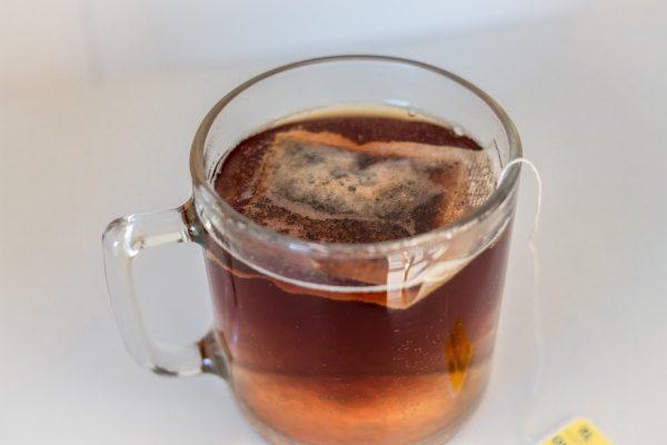 Plano picado de una taza de vidrio transparente llena de té infusionado, donde se observa en la parte superior un saquito de té flotando sumergido cerca de la superficie.