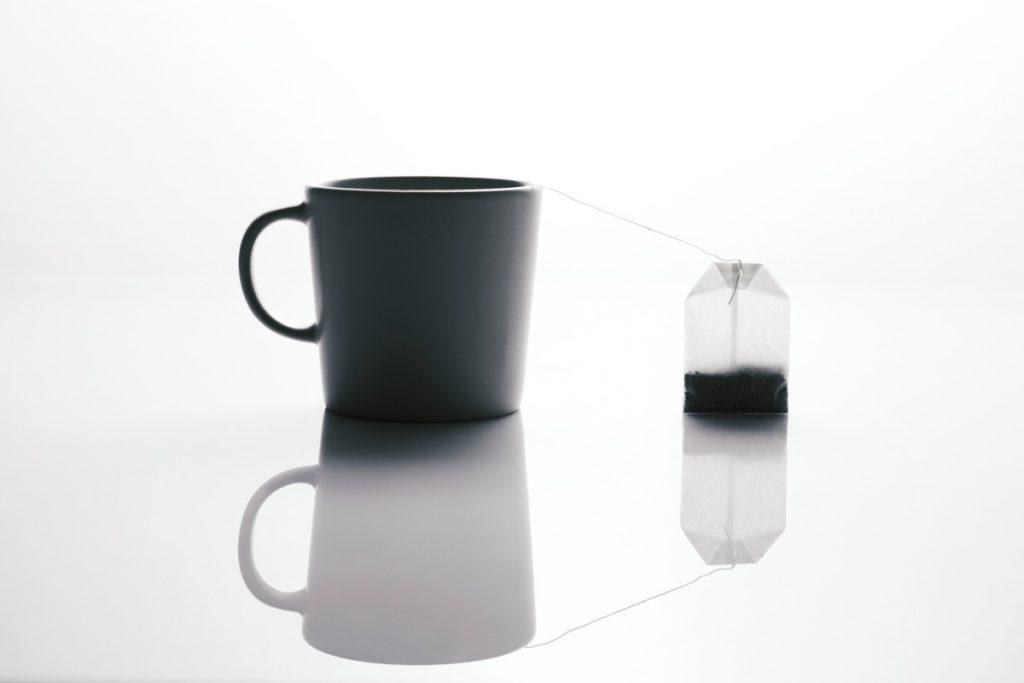 Fotografía minimalista en tono de silueta sobre fondo blanco brillante que muestra una taza de cerámica oscura a la izquierda y una bolsa de té llena de hebras suspendida a la derecha por su hilo, reflejándose ambas en una superficie pulida.