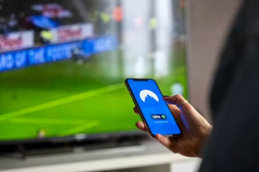 Persona sosteniendo un teléfono móvil con una aplicación de VPN abierta frente a un televisor que transmite un partido de fútbol